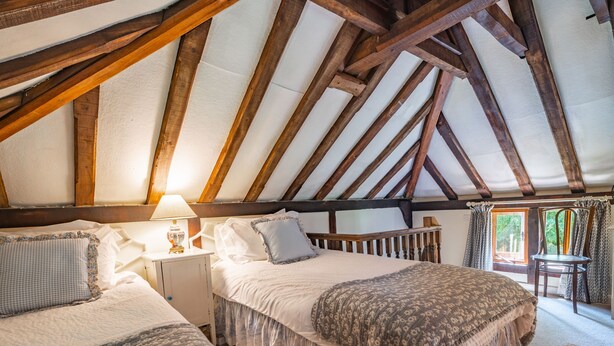 Une chambre avec un plafond en pente, des poutres en bois, deux lits avec des têtes de lit assorties, une table de chevet avec une lampe et une fenêtre avec des rideaux.