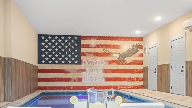 Zona de piscina con un mural de la bandera estadounidense y un águila, una fuente de agua y una mesa con bebidas.