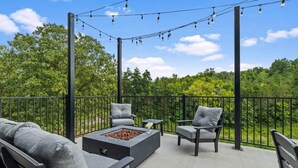 Un patio exterior con una chimenea, asientos e iluminación de guirnaldas, frente a un fondo de árboles.