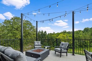 Eine Terrasse im Freien mit Feuerstelle, Sitzgelegenheiten und Lichterketten vor einer Baumkulisse.