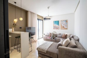 Una sala de estar moderna con un sofá, una mesa de centro y un televisor. Hay una zona de comedor con una mesa y sillas. La habitación tiene ventanas amplias y un ventilador de techo.