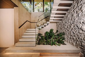 A modern staircase with a wooden finish, a stone wall, and a view of greenery outside. - New · 180° Views – River & National Park · Infinity Pool · 3 min to the Beach (Tamarindo)