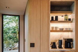 A modern kitchen with built-in wooden cabinets, a coffee station, and a view of a garden through a large window. - New · 180° Views – River & National Park · Infinity Pool · 3 min to the Beach (Tamarindo)