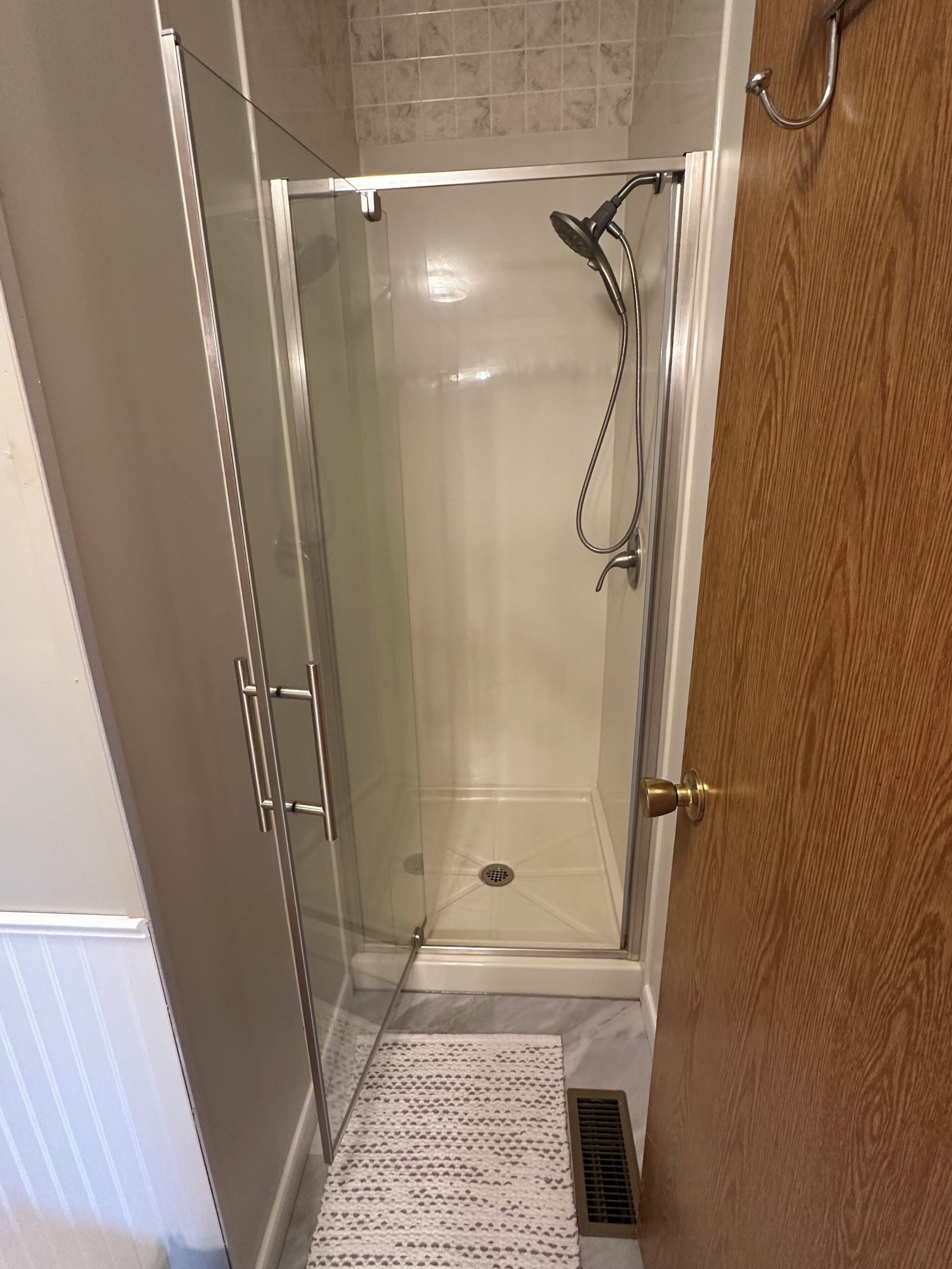 A glass-enclosed shower with a showerhead and a drain, a wooden door, and a tiled floor.