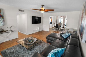 A modern living room with a leather sofa, a coffee table with decorative items, a flat-screen TV mounted on the wall, and a ceiling fan. - Beautiful 4-bedroom House With Heated Swimming Pool & Spa (Fort Lauderdale)