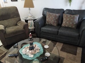 Ein Wohnzimmer mit Glastisch, einer Couch, einem Sessel und einem kleinen Tisch mit Lampe.