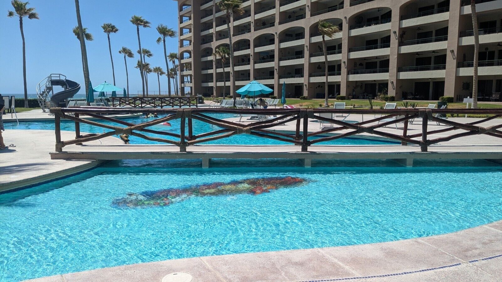 A swimming pool with a diving board, surrounded by palm trees and a multi-story building in the background.