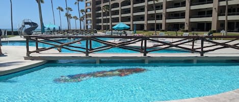 A swimming pool with a diving board, surrounded by palm trees and a multi-story building in the background.