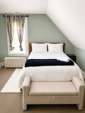 A bedroom with a bed, a nightstand, a radiator, a window with curtains, and a carpeted floor.