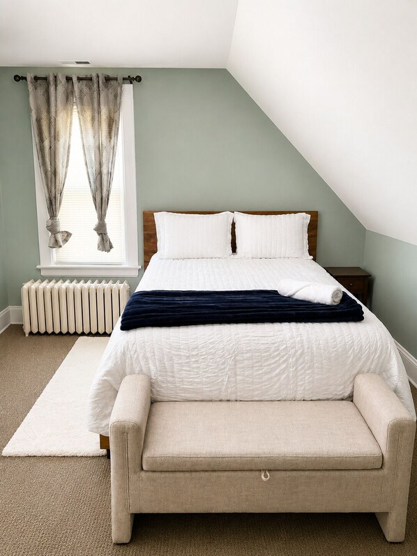 Une chambre à coucher comprenant un lit, une table de chevet, un radiateur, une fenêtre avec des rideaux et un sol recouvert d’une moquette.