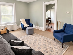 A living room with a sofa, two armchairs, a rug, a radiator, and a dining area in the background.