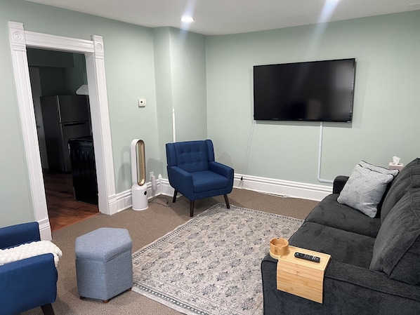 A living room with a flat-screen TV mounted on the wall, a blue armchair, a grey ottoman, and a grey sofa with a remote on a wooden tray.