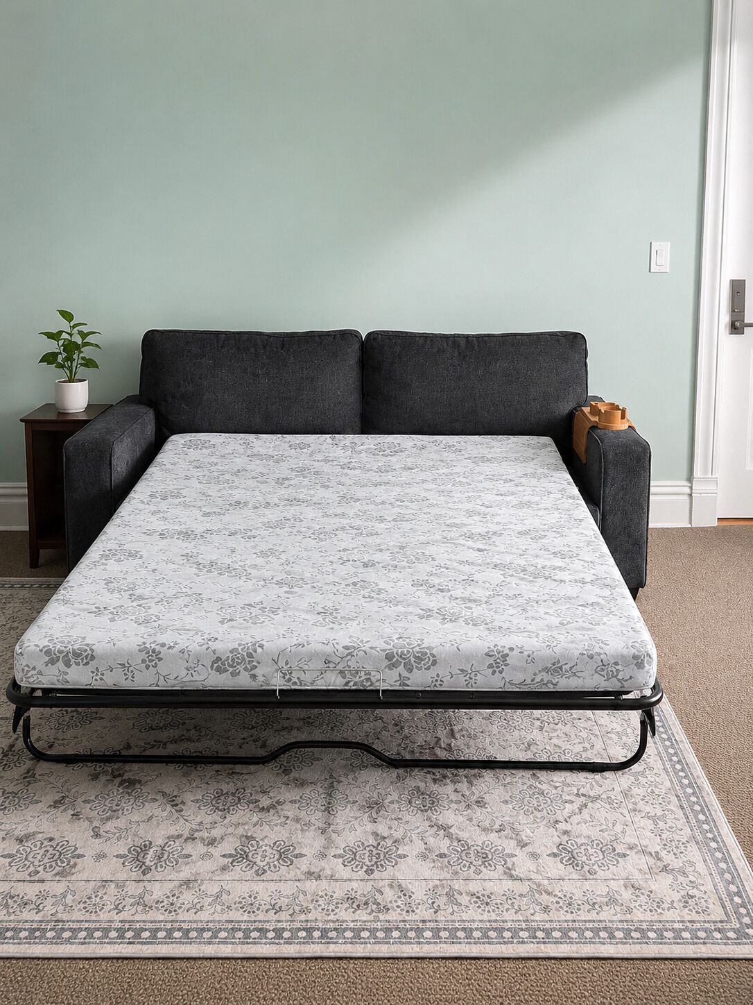 A bed with a floral mattress, a dark sofa, a small table with a plant, and a door in the background.