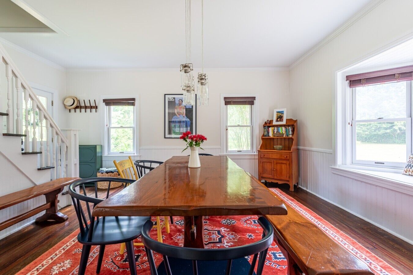 A dining room with a wooden table, chairs, a vase with flowers, a bookshelf, and a staircase.