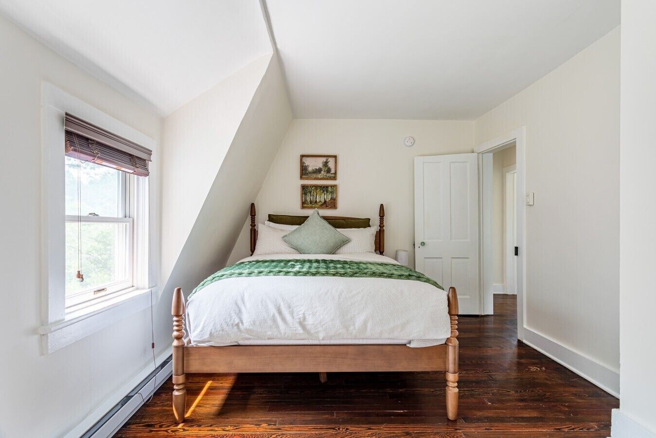 Ein Schlafzimmer mit einem hölzernen Bett, weißen Wänden, einem Fenster mit Jalousien und einer Tür zu einem anderen Raum.