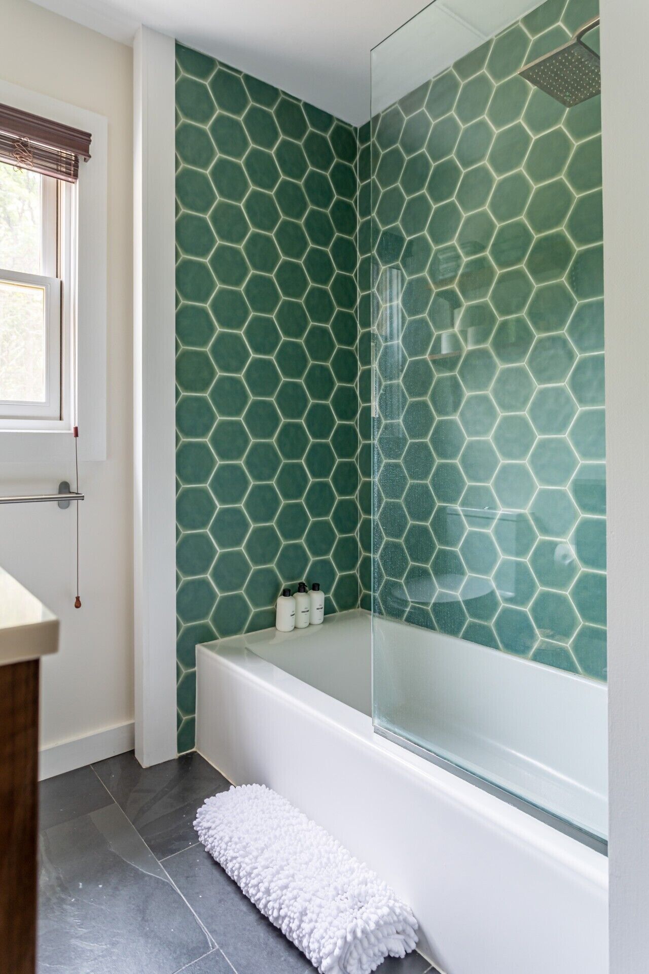 Une salle de bain avec un carrelage à motifs hexagonaux, une douche en verre, une baignoire blanche et un tapis de bain blanc.