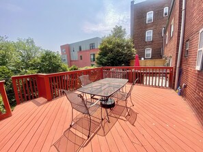 Une terrasse en bois avec une table et des chaises, entourée d’une rambarde rouge, et des bâtiments en arrière-plan.