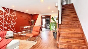 A modern living room with a wooden staircase, a dining area, and a kitchen in the background. - Direct PATH & Bus to NYC | MetLife 20 min | Parking + Deck (Jersey City)