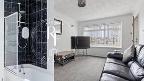 A modern bathroom with a shower, a bathtub, and a toilet. There is a television in the living room area, a window with blinds, and a leather sofa. - The Den at Teal Court (Peterhead)