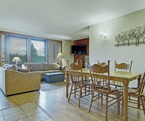 Ein Wohnzimmer mit einer Couch, einem Esstisch mit Stühlen, einem an der Wand befestigten Fernseher und einem Fenster mit Jalousien.