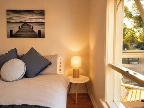 Ein Schlafzimmer mit einem Bett, Kissen, einer Nachttischlampe, einem Bild an der Wand und Blick durch ein Fenster.
