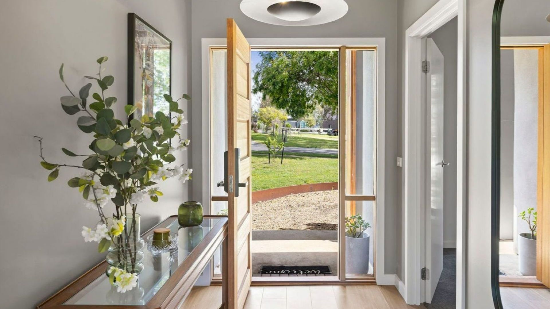A hallway with a glass door leading to an outdoor area, a wooden bench with a vase of flowers, and a framed picture on the wall.