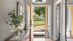 Un pasillo con puerta de cristal que da a un espacio exterior, un banco de madera con un jarrón de flores y un cuadro en la pared.