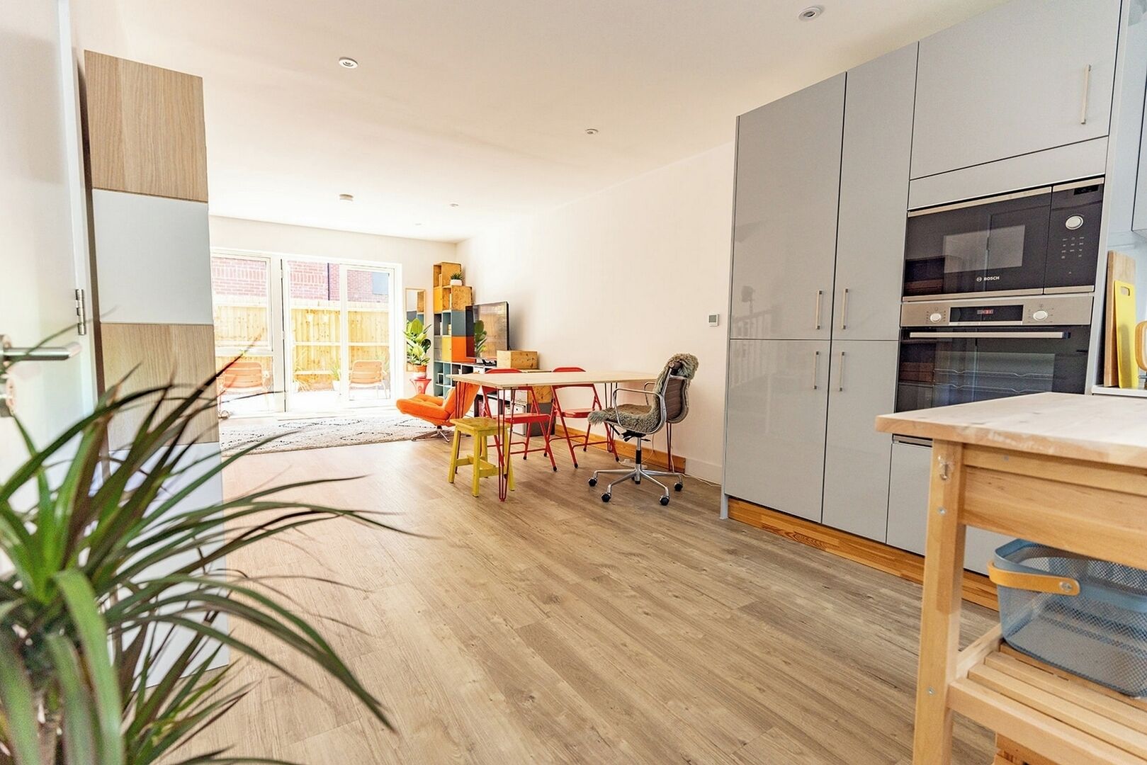 Een moderne keuken met grijze kastjes, een Bosch oven, en een eethoek met een houten tafel en kleurrijke stoelen.