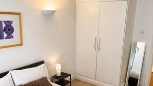 Een slaapkamer met een bed, een garderobe, een nachtkastje, een lamp, een spiegel en een lijst met een foto aan de muur.