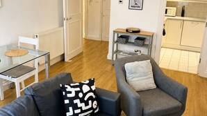 Een moderne woonkamer met een bank, fauteuil en een glazen salontafel. In de achtergrond is een keuken te zien met kastjes en een magnetron.