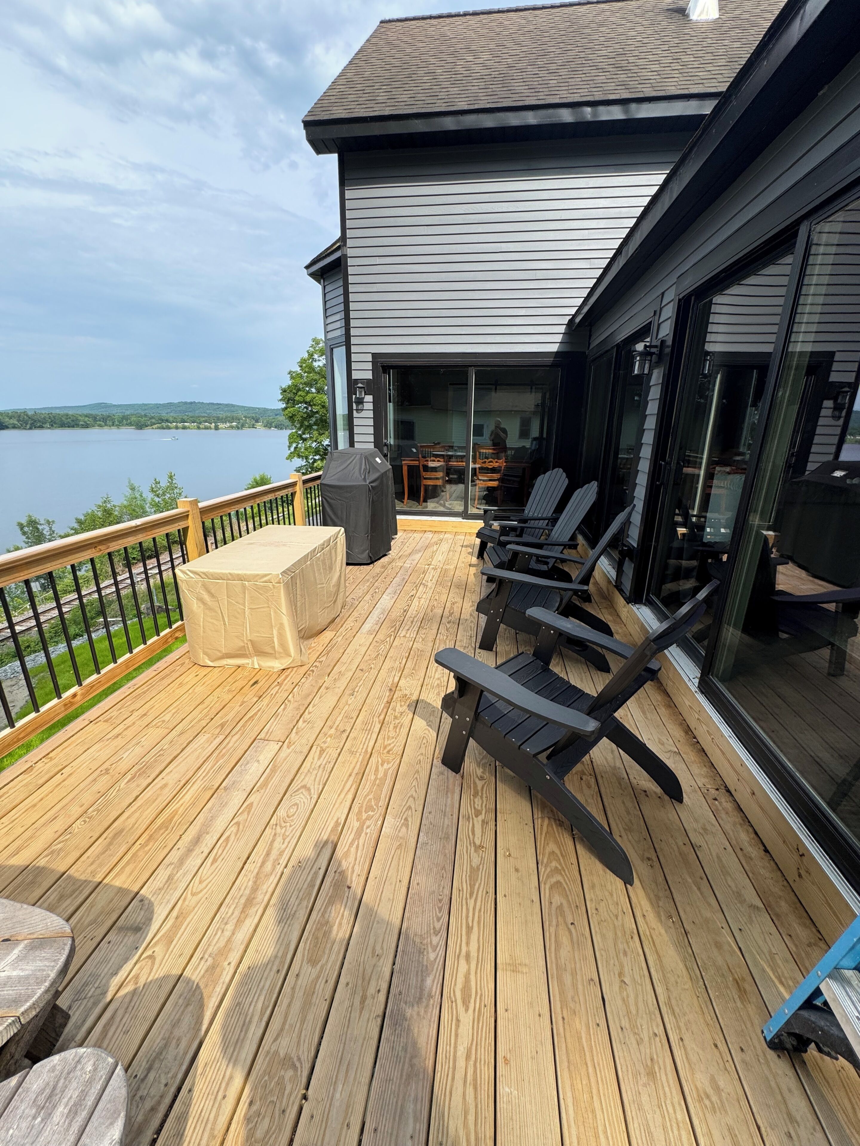 Una terraza de madera con mobiliario exterior, una parrilla cubierta y vistas a un lago.