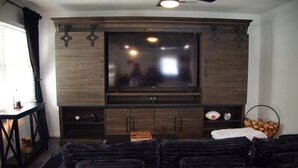 A living room with a large flat-screen TV mounted on a wooden cabinet, a ceiling fan, and a fireplace with a log basket. - 3BR Downtown Cleveland Home Near Stadiums Free Parking (Cleveland)