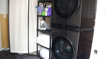 Un espace buanderie compact comprenant deux machines à laver, une étagère avec des produits de nettoyage et un rideau blanc.