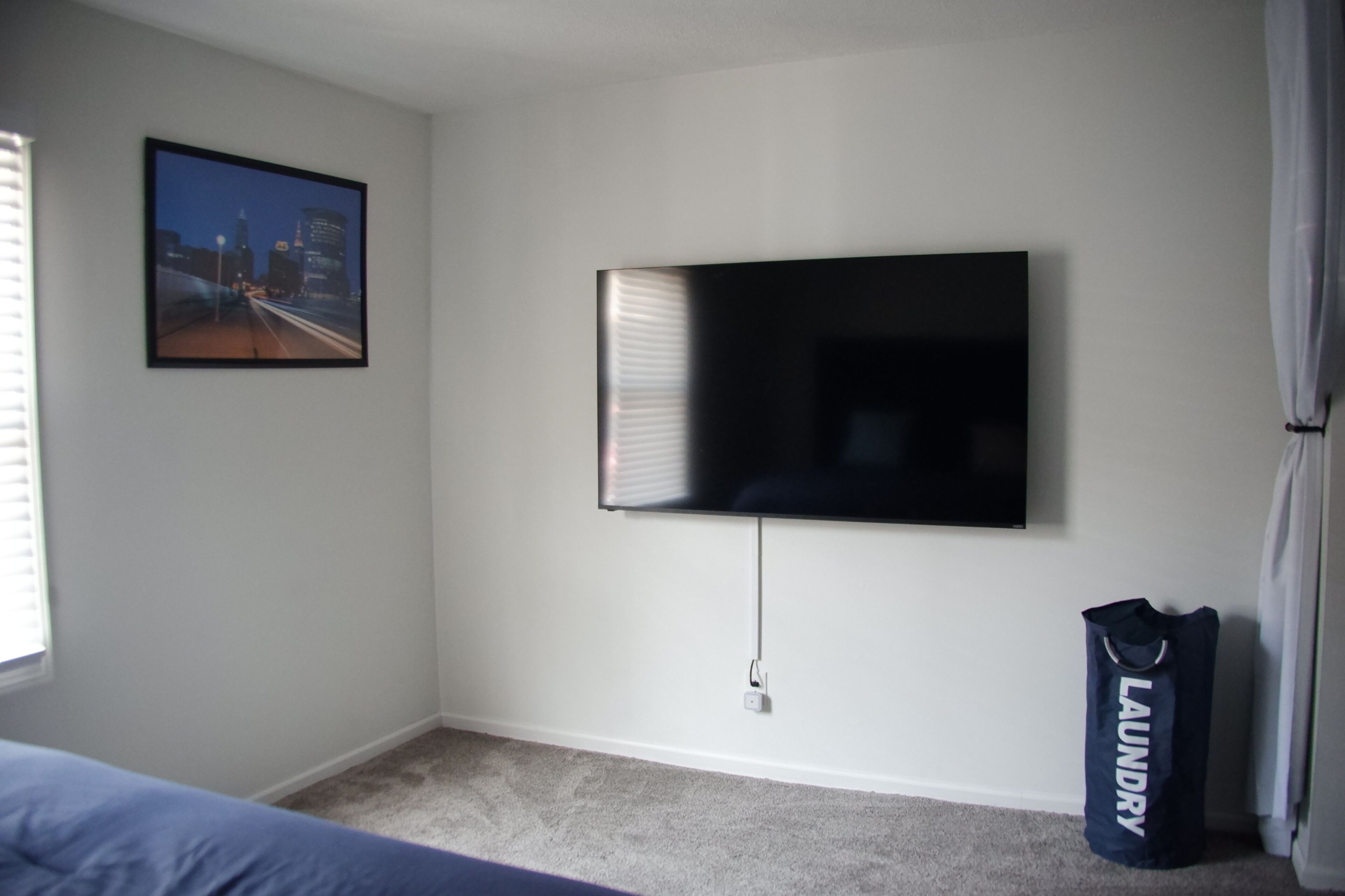 Une chambre dotée d’un téléviseur à écran plat fixé au mur, d’une fenêtre avec des stores et d’un sac à linge posé sur le sol.