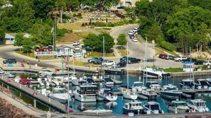 A marina with multiple boats docked, a parking area with cars, and a grassy area with people.