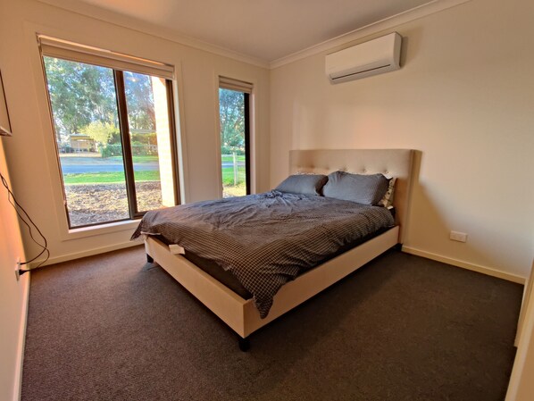 A bedroom with a bed, large windows, an air conditioning unit, and carpeted floor.