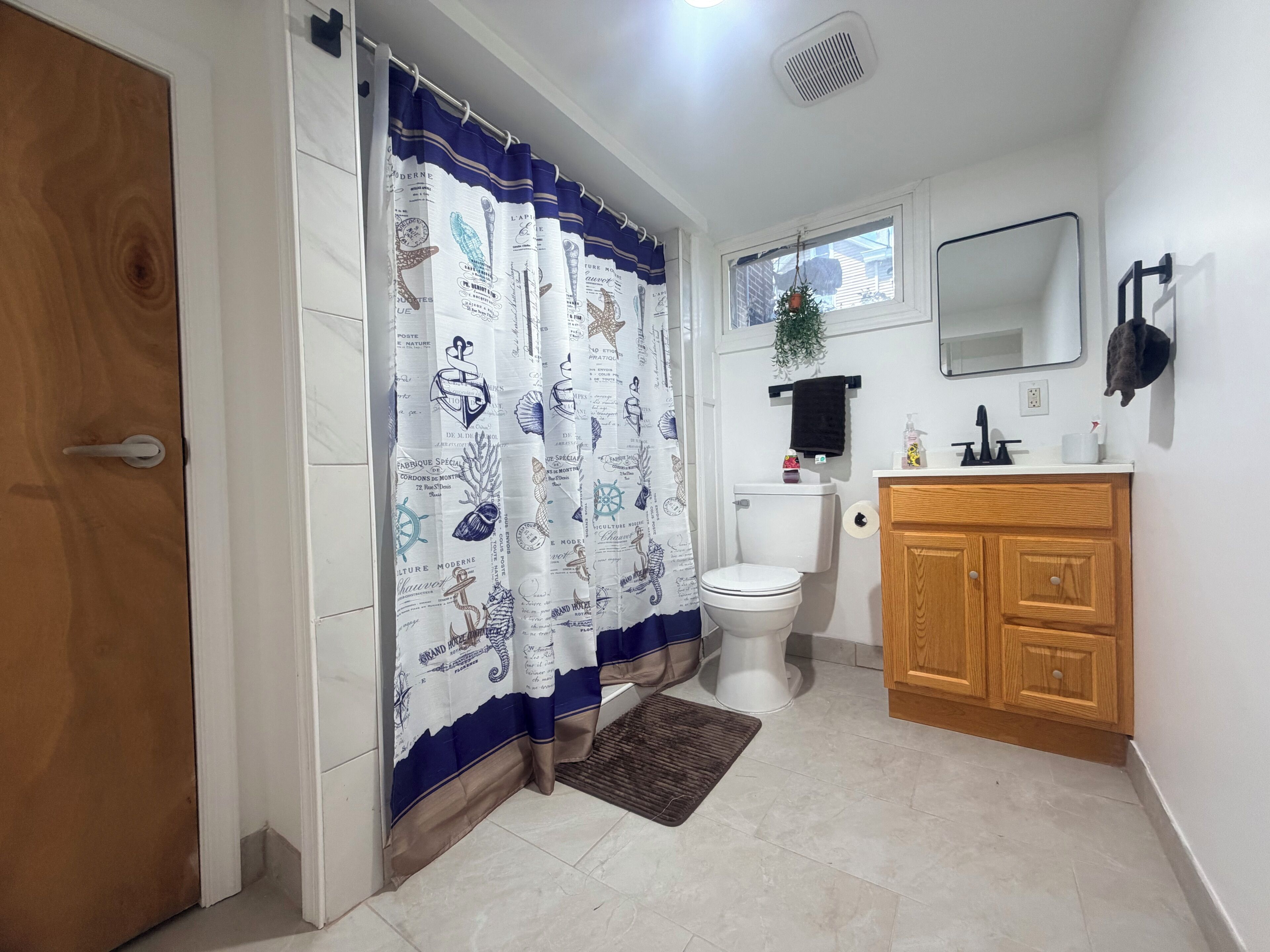 A bathroom with a white toilet, a wooden cabinet, a mirror, and a shower curtain with nautical designs.