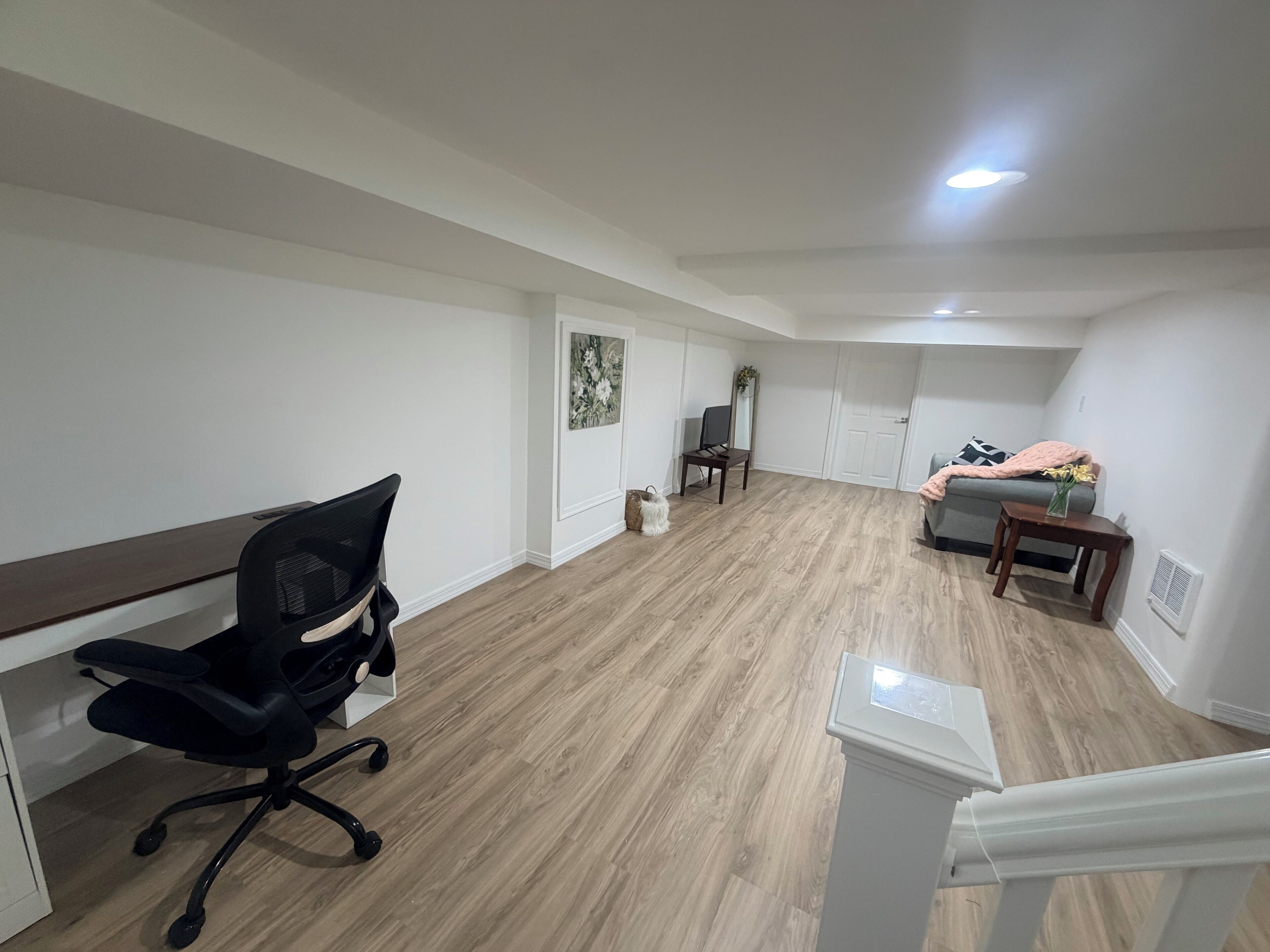 A spacious room with a wooden floor, a desk with a chair, a bed with a blanket, and a small table with a vase.