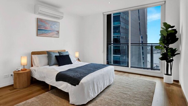 Quarto moderno com uma cama grande, mesa de cabeceira, abajur, janela com vista para edifícios e um vaso com planta.