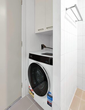 A compact laundry room with a front-loading washing machine, a small cabinet above it, and a towel rack on the wall. - Stunning L34 views | Pool, Sauna, Gym | Sth Cross (Melbourne)