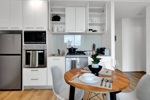 A modern kitchen with white cabinetry, a built-in oven, microwave, and a dining area with a wooden table and chairs. - Stunning L34 views | Pool, Sauna, Gym | Sth Cross (Melbourne)