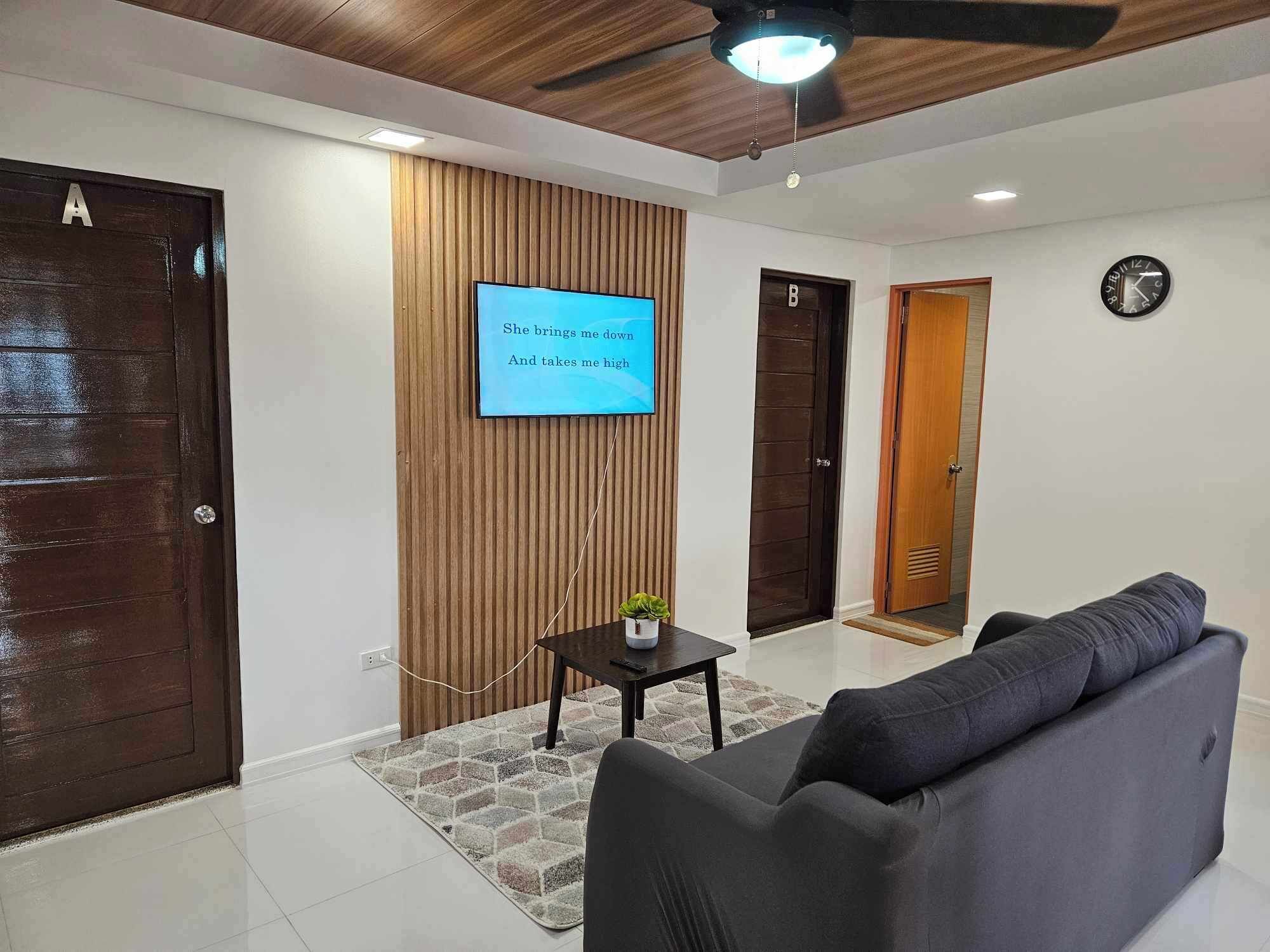 A modern living room with a flat-screen TV mounted on a wooden wall, a sofa, a small table with a plant, and a ceiling fan.