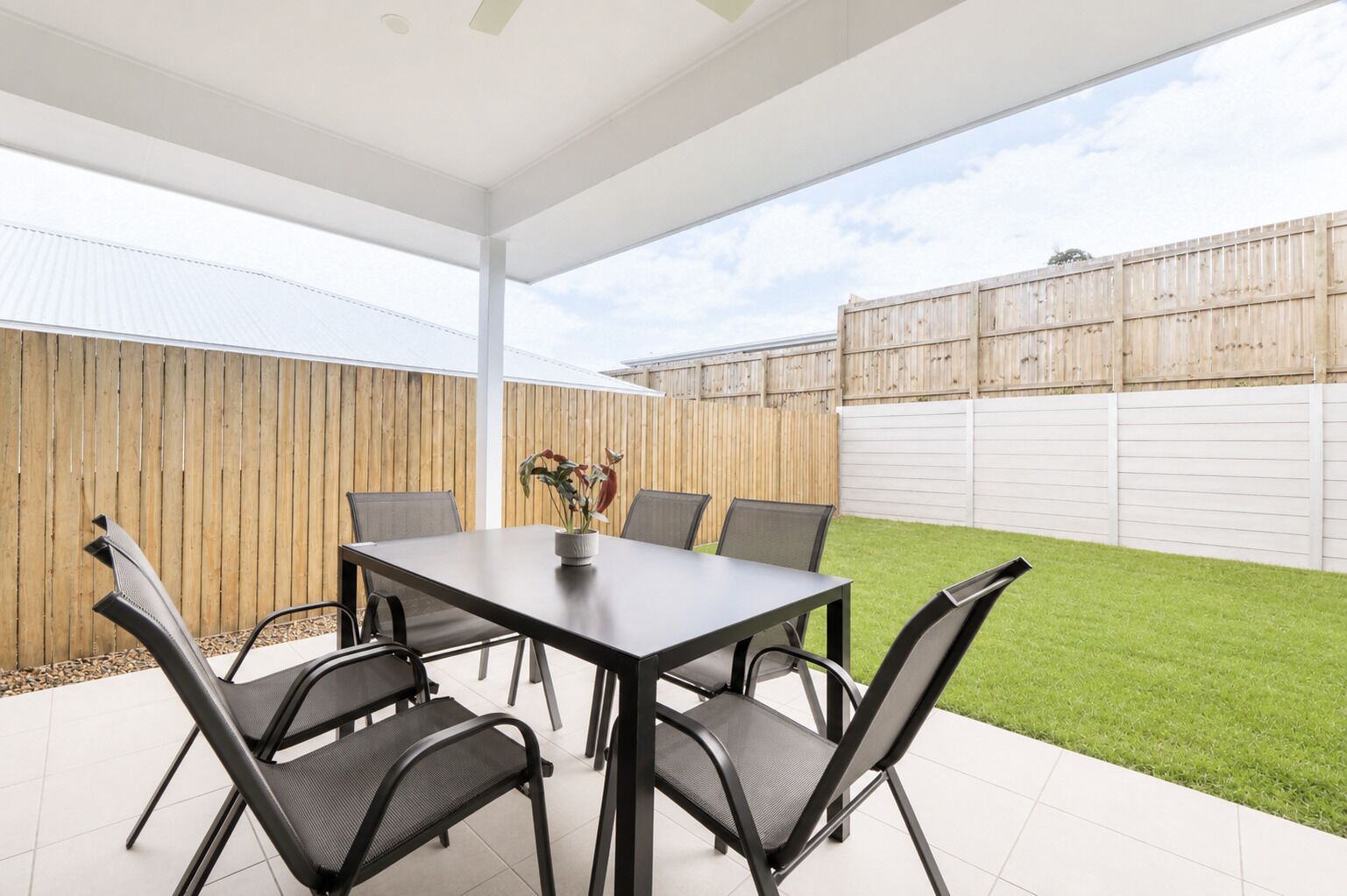 A covered patio area with a table and chairs, a potted plant, and a wooden fence in the background.