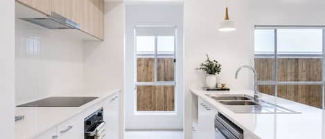 A modern kitchen with white cabinetry, a built-in oven, a dishwasher, and a window with a wooden view.