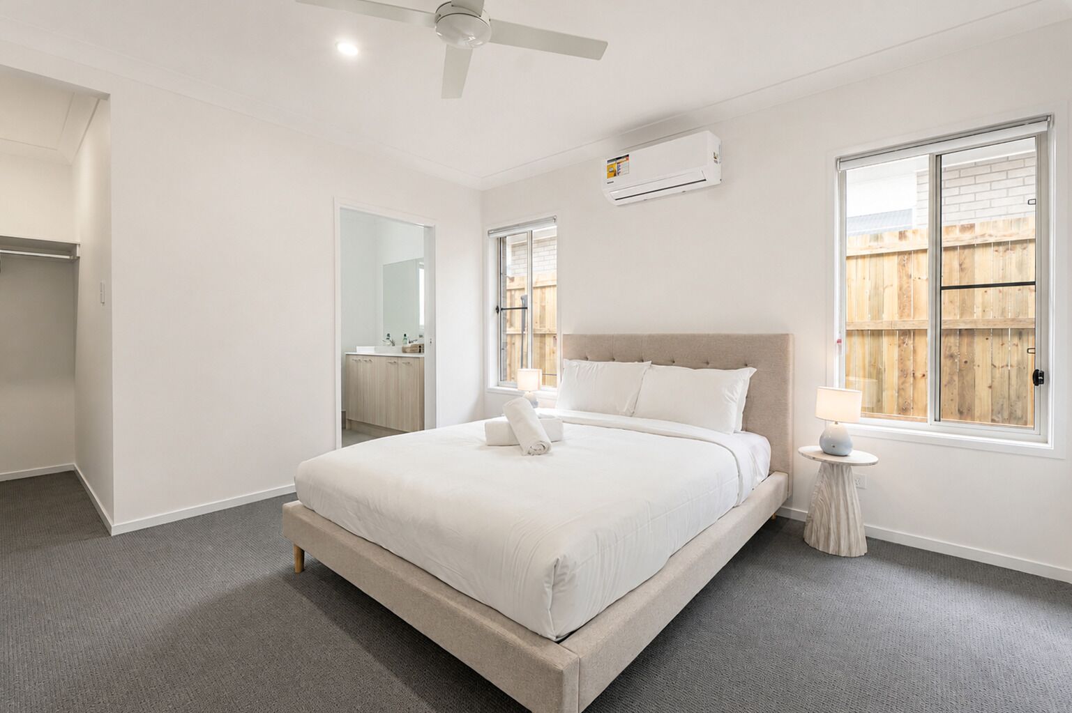 A bedroom with a bed, a bedside table, a ceiling fan, an air conditioning unit, a window, and a view of a wooden fence.