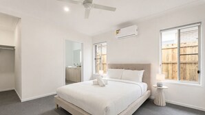 Une chambre à coucher comprenant un lit, une table de chevet, un ventilateur de plafond, un appareil de climatisation, une fenêtre et une vue sur une clôture en bois.