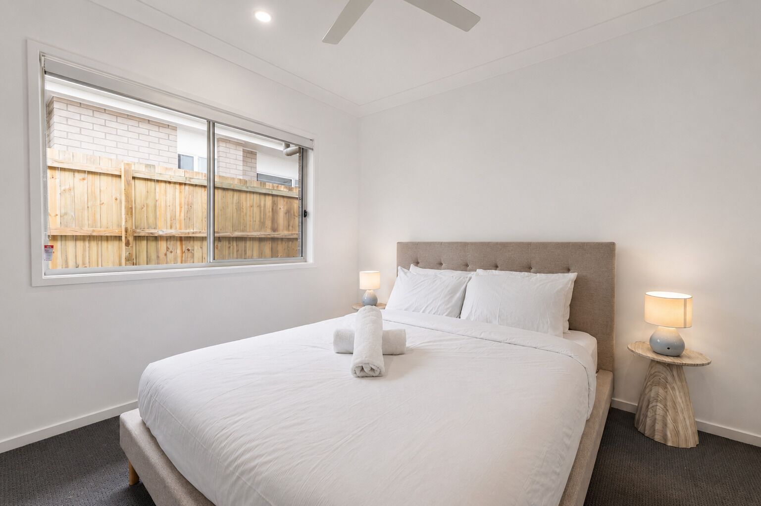 A bedroom with a large bed, two bedside lamps, a window with a view of a wooden fence, and a ceiling fan.