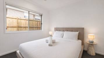A bedroom with a large bed, two bedside lamps, a window with a view of a wooden fence, and a ceiling fan.
