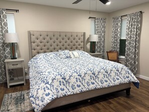 A bedroom with a tufted headboard, a floral bedspread, two bedside tables with lamps, a wooden floor, and a window with patterned curtains.