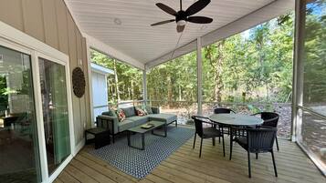 A covered outdoor area with a ceiling fan, a sofa set, a dining table with chairs, and a view of a forested area.